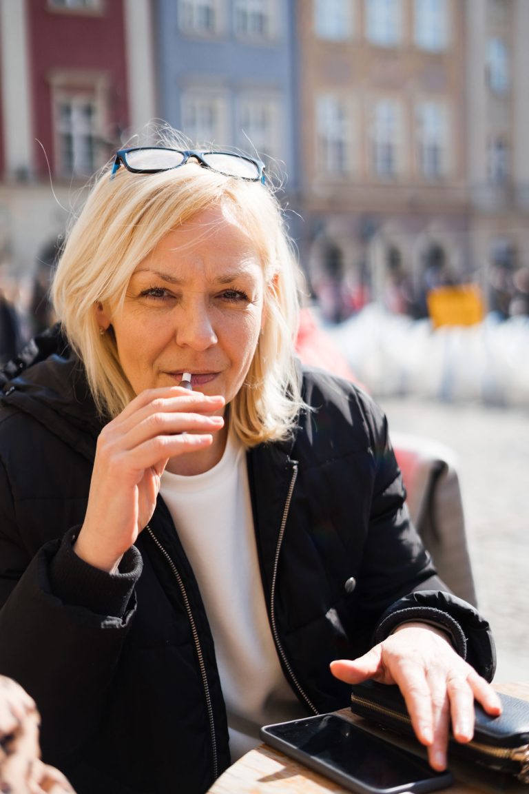 Middle aged blonde European woman smoking heated tabacco e-cigarette outdoors on the street. Alternative smoking way.